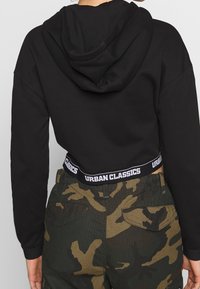 Svart cropped hoodie med huva, kombinerad med kamouflage cargo-byxor med en svart elastisk midja märkt "URBAN CLASSICS."