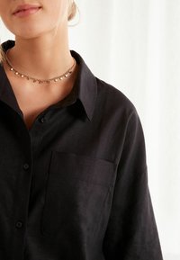 Czarna koszula z guzikami, wykonana z teksturowanej tkaniny, z kieszonką na piersi oraz klasycznym kołnierzykiem. Noszona z warstwowym metalicznym naszyjnikiem choker.