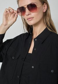 Melna pogāžama jaka ar divām krūšu kabatām, gludu virsmu un apkakli. Modelis valkā tonētas saulesbrilles ar zelta ietvariem.