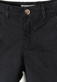 Gros plan sur la ceinture d'un pantalon noir avec une fermeture par bouton, des passants de ceinture et des détails de coutures visibles.