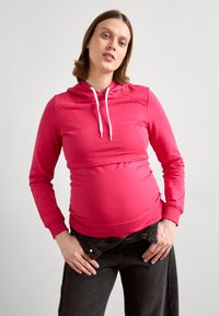 Rožinė ilgomis rankovėmis kapučino striukė su kengūros kišene ir baltomis virvutėmis, sukurta su prigludusia viršutine dalimi ir reguliuojama apatine dalimi patogumui.