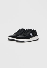 Czarne niskie sneakersy z białymi grubymi podeszwami i białym haftowanym logo po bokach, wyeksponowane na jednolitym jasnoszarym tle.