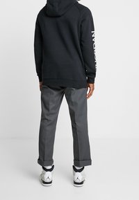 Felpa nera con logo bianco sulla manica, abbinata a pantaloni grigi chiari. Sneakers bianche con accenti grigi e polsini risvoltati.