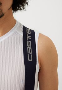 Maglia da ciclismo bianca in rete con bretelle blu navy che presenta il logo "Castelli" in grigio. Primo piano della spalla e del collo, evidenziando la texture del materiale.