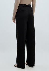 Zwarte wijde broek met een getailleerde pasvorm, voorzien van twee achterzakken en een zachte textuur. G gedragen met een lichtgekleurde langemouwen top.