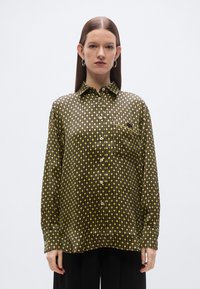 Melna pogājama krekla ar ģeometrisku dzeltenu rakstu, kas ir apģērbta ar apkakli, garām piedurknēm un krūšu kabatu. Gluda auduma tekstūra.