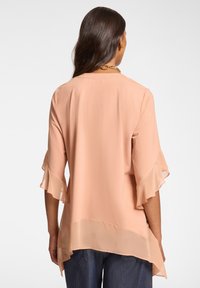 Šifonová blůza v broskvové barvě s rozšířenými rukávy, která má delší zadní lem a průhlednou texturu. Jednoduchý kulatý výstřih, lehký design.
