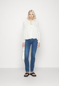 Blusa branca com decote em laço, pontos texturizados subtis, mangas compridas, combinada com jeans skinny azuis e sandálias pretas de slip-on. Bolsa branca.