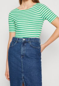 Nainen, joka on pukeutunut vihreä-valkoisiin vaakasuoriin raitapaitoihin, jotka on tavanomaisesti sisätetty korkeavyötäröiseen siniseen farkkuhameeseen, jossa on edessä halkio.