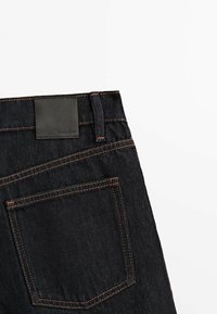 Black denim jeans with contrasting orange stitching, featuring a leather patch at the waistband and a single back pocket.