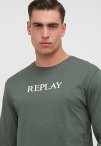Zeleni dukserica dugih rukava od pamuka, s bijelim "REPLAY" logotipom na prsima. Standardni dizajn s okruglim izrezom.