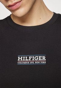 T-shirt di cotone nera con un design grafico di testo bianco e blu che recita "HILFIGER COLUMBUS AVE. NEW YORK." Maniche corte e collo rotondo.