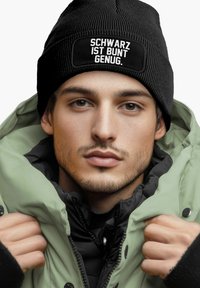 Black knitted beanie with white text, paired with a green oversized jacket, featuring a high collar and black inner lining.