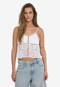 White cropped top with lace trim, featuring a tie front and layered design. Paired with light blue high-waisted jeans.