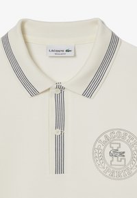 Krém színű pólóing, texturált felülettel; sötétkék csíkos gallérral, két gombbal és kör alakú Lacoste logóval rendelkezik.