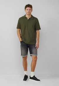 Oliwkowa koszulka polo z kołnierzykiem, zestawiona z podwiniętymi ciemnoniebieskimi szortami dżinsowymi. Czarnie sneakersy i białe skarpetki dopełniają stylizację.
