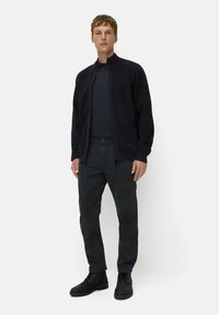 Donkere, ritssluiting jack met een hoge kraag, gecombineerd met een bijpassend shirt en donkere broek. Robuuste zwarte laarzen maken de look compleet.