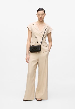 Vrouw draagt een beige mouwloze dubbelrijige blazer en wijde pantalon, met een zwarte crossbodytas, staand tegen een witte achtergrond.