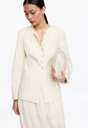 Kvinde med mørkt hår iført en cremefarvet jakke med knapper og en lys nederdel, der holder en matchende cremefarvet snøresæk mod en hvid baggrund.