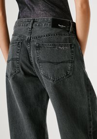 Pelēki džinsi ir ar augstu jostasvietu, lieliem aizmugures kabatām un smalku zīmolu izstrādājumu jostasvietā. Audumam ir teksturēts apdare.