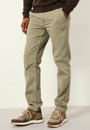 Man wearing slim-fit olive green pants and brown casual sneakers, standing with one hand in pocket against a plain background.