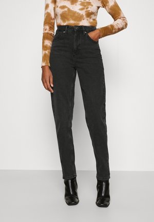 Persona che indossa jeans neri a vita alta, stivaletti neri e una maglia a maniche lunghe tie-dye marrone e bianca, in piedi con una mano in tasca.