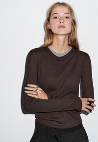 Braunes Langarmoberteil mit grauem Ausschnitt, glattes Material, figurbetonte Silhouette. Getragen mit schwarzen Hosen, accessoriert mit mehreren Ringen.