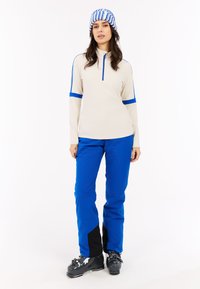 Beige zip-up top met blauwe accenten en lange mouwen, gecombineerd met blauwe broek en zwarte accenten, gedragen met zwarte skiboots.