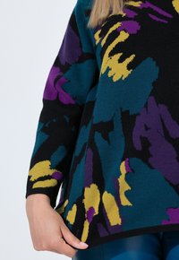 Frau trägt einen langärmligen Pullover mit abstraktem Muster in Gelb, Lila, Türkis und Schwarz und hält den Saum über blaue Hosen vor einem weißen Hintergrund.