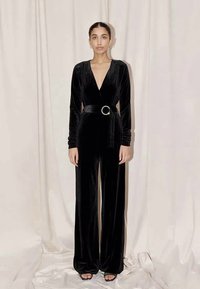 Black velvet jumpsuit with long sleeves, deep V-neck, and wide legs. Features a belt with a circular jeweled buckle for added detail.