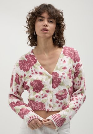 Donna con capelli ricci che indossa orecchini a cerchio e un cardigan bianco a vita corta con grandi motivi floreali rosa, abbottonato davanti.