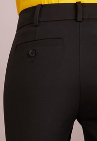 Černé kalhoty na míru s hladkou texturou, s zadní kapsou na knoflík a poutky na opasek v pase.