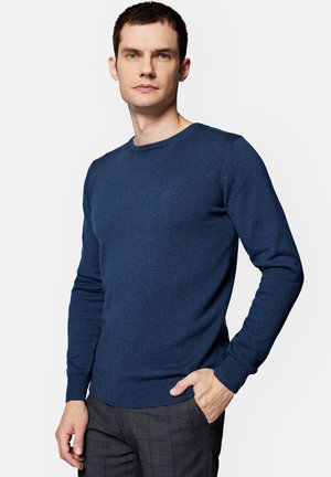 Granatowy sweter z dzianiny z okrągłym dekoltem, prążkowanymi mankietami i dołem. Ozdobiony małym, subtelnym logo na piersi. Miękka tekstura.