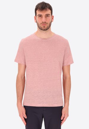 Uomo in piedi di fronte, indossa una maglietta tinta unita rosa chiaro a maniche corte e pantaloni scuri su uno sfondo semplice.