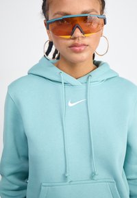 Hoodie vert menthe avec une poche avant et un logo Nike blanc. Le modèle porte des lunettes de soleil teintées d'orange avec des accents bleus et de grandes boucles d'oreilles en argent.