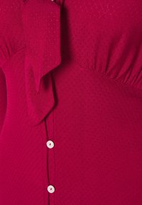 Blouse rouge texturée avec un détail nœud papillon au niveau du col et de petits boutons blancs le long de la couture latérale.