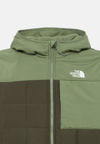 Doudoune verte avec capuche, texture matelassée, panneaux contrastants en vert foncé et poche sur la poitrine gauche avec logo.