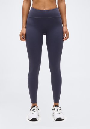 Vista frontale di una persona che indossa leggings grigio scuro a vita alta e scarpe da corsa bianche, in piedi contro uno sfondo chiaro e uniforme.