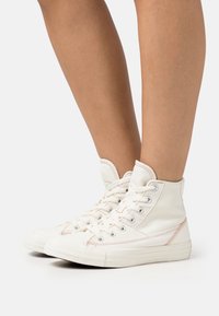 Białe płócienne sneakersy w stylu high-top z kontrastującymi przeszyciami, okrągłym noskiem i sznurowanym zapięciem. Gładka tekstura i gumowa podeszwa.