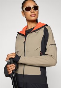 Lichte, beige jas met een zwarte accent en een feloranje fleece-voering capuchon. Gecombineerd met wandelstokken, met een ceintuurzak.