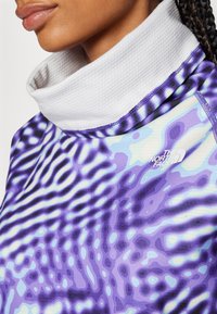 Pullover ülikond, millel on kõrge valge krae, ja lilat ning sinist abstraktset mustrit. Valmistatud sileda, veniva kanga abil, millel on õrn tekstuur.