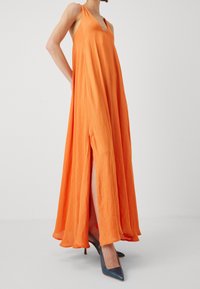Robe maxi sans manches orange avec un décolleté en V, dotée de fentes sur les côtés et d'un design fluide, associée à des escarpins bleus à bout pointu.