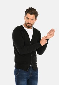 Cardigan nero con maniche lunghe, bottoni sul davanti e polsini a coste; indossato sopra una camicia bianca, abbinato a jeans blu.