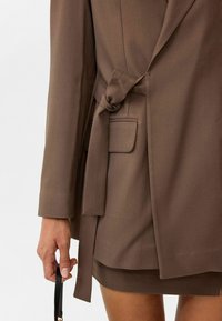 Bruine blazer met een gebonden taille, zachte textuur, inkepingen op de revers en een zijzak. Hand die een zwarte band vasthoudt, met een gelaagde stof eronder.