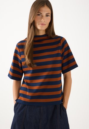 Frau mit langen braunen Haaren, die ein marineblau-braun gestreiftes Kurzarmoberteil und dunkle Jeans trägt, mit den Händen in den Taschen.