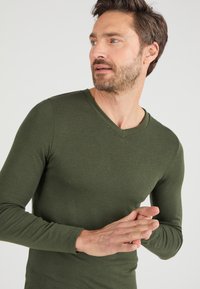 Olijfgroene V-hals shirt met lange mouwen gemaakt van een zachte, gestructureerde stof. Aansluitend ontwerp met omslagmouwen en minimale naden.