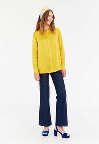 Chemise à manches longues jaune avec des boutons, jean évasé foncé et sandales à plateforme bleues. Le modèle porte un bandeau à motifs. Fond blanc uni.