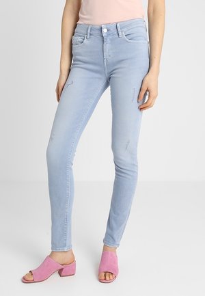 Kvinde iført lyseblå, tætsiddende jeans med let slid samt lyserøde blokhælssandaler med åbne tæer foran en ensfarvet baggrund.