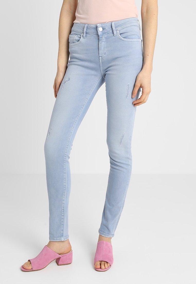 Femme portant un jean slim bleu clair avec de légères déchirures et des sandales à talons carrés ouvertes roses sur un fond uni.