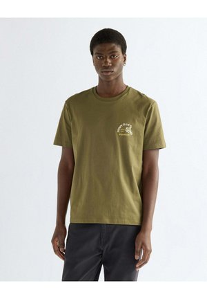 Junger Mann steht, trägt ein olivgrünes Wrangler Denim Club T-Shirt und schwarze Hose vor einem schlichten hellen Hintergrund.